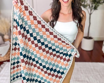 May include: A rectangular crochet blanket featuring a repeating granny square pattern in a colour palette of cream, dark teal, muted orange, and deep burgundy. The blanket has a cream-coloured border and a small brown rectangular tag at the corner.