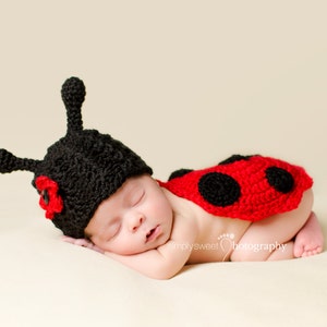 May include: A sleeping baby wearing a black and red ladybug costume. The costume is crocheted and has a black hat with red accents.