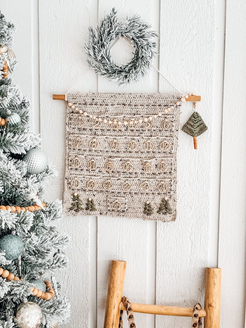 May include: A handmade crochet advent calendar with wooden numbers and small Christmas tree accents. The calendar is beige and hangs from a wooden dowel. A small green crochet Christmas tree hangs on the right side. A frosted Christmas tree and wreath are in the background.