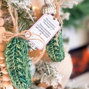 May include: Two green crocheted pickle ornaments hanging from twine strings. A white rectangular tag with red scalloped edges is attached, reading: Hide this pickle in your tree and let the chaos begin! Whoever finds it first on Christmas morning gets a sprinkle of luck.