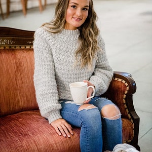 Può includere: Un maglione in maglia a maniche lunghe color grigio chiaro con un motivo testurizzato a pallini sulla parte superiore del petto e sulle maniche, abbinato a jeans blu strappati. Il maglione presenta un girocollo a coste e polsini a coste.