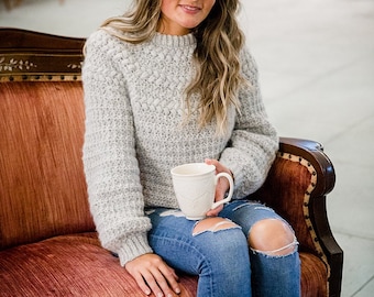 May include: A light gray long-sleeved knit sweater with a textured bobble pattern on the upper chest and sleeves, paired with blue distressed denim jeans. The sweater features a ribbed crew neckline and ribbed cuffs at the wrists and waist.