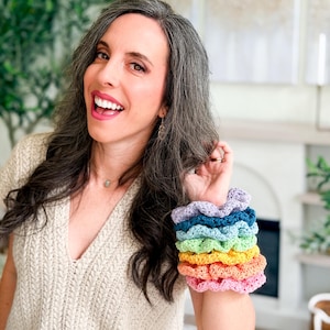 May include: A stack of six crocheted hair scrunchies in a rainbow color gradient, featuring shades of light purple, dark blue, teal, light green, yellow, orange, and pink, arranged in a vertical column on a wrist.