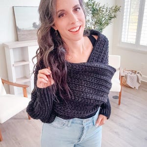 A woman stands indoors wearing a black crochet shawl and jeans, smiling at the camera. A chair, a small shelf with unique jewellery, and a framed picture are in the background.