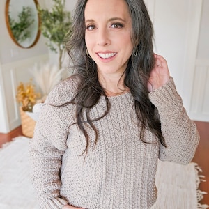 A woman with long hair and a gray knit sweater smiles at the camera in a well-lit room with a round mirror, plants, and unique jewellery displayed in the background.