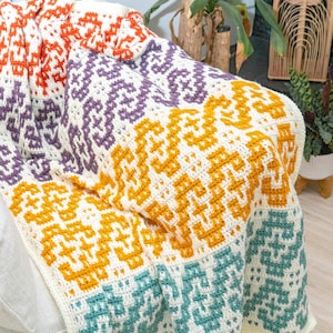 Puede incluir: Una manta de ganchillo multicolor con un patr&oacute;n geom&eacute;trico en tonos naranja, morado, amarillo y turquesa. La manta est&aacute; colocada sobre un sof&aacute; blanco.