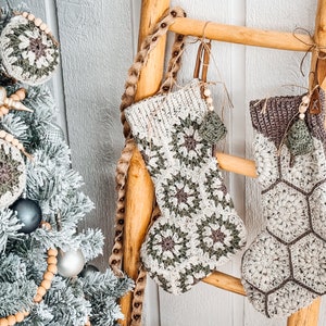 May include: Two handmade crochet Christmas stockings hanging on a wooden ladder. One stocking features a cream and sage green granny square pattern, while the other has a cream and brown hexagonal motif. Both include wooden beads and small tree charms.