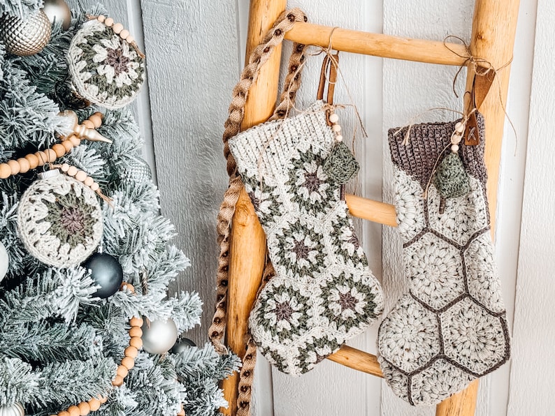 May include: Two handmade crochet Christmas stockings hanging on a wooden ladder. One stocking features a cream and sage green granny square pattern, while the other has a cream and brown hexagonal motif. Both include wooden beads and small tree charms.