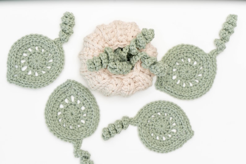 Cream crochet pumpkin surrounded by four sage green leaf coasters with spiral stems on a white surface