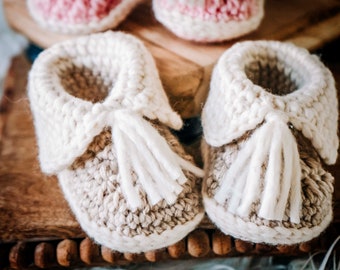 Könnte beinhalten: Ein Paar beige gestrickte Babyschuhe mit weißen Quasten. Die Schuhe haben einen weißen gestrickten Saum am oberen Rand.