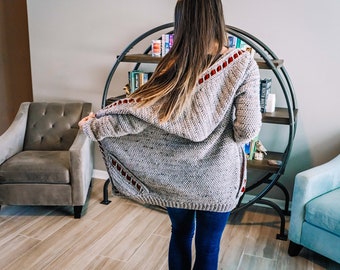 May include: A light gray speckled knit hooded cardigan featuring long sleeves and dark red rectangular cut-out accents along the hood and front panels. The garment is worn over dark blue denim jeans, showcasing a textured yarn construction.