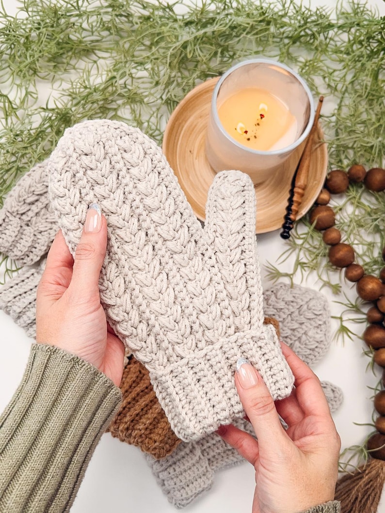 May include: A pair of beige crocheted mittens with a cable knit pattern. The mittens are held in a person's hands, with one mitten being held up to show the detail of the knit.