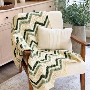 Puede incluir: Una manta de ganchillo con un patrón de chevrón en rayas alternas de color crema, verde salvia y verde bosque oscuro. La manta está colocada sobre un sillón de color crema que también sostiene un pequeño cojín rectangular de color crema.