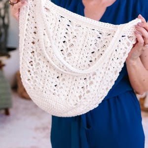 Puede incluir: Bolso de crochet blanco roto con tejido abierto y correa para el hombro. El bolso tiene forma redondeada y superficie texturizada. El bolso lo sostiene una persona con un mono azul.