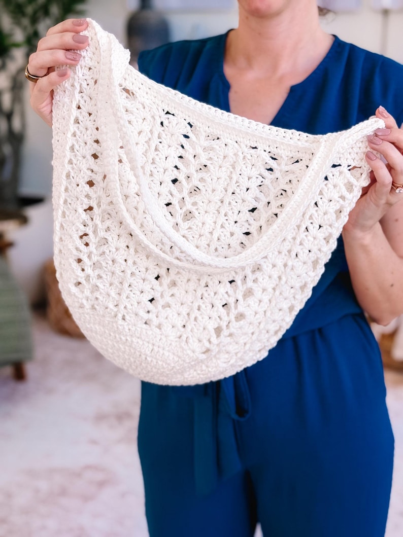 Puede incluir: Bolso de crochet blanco roto con tejido abierto y correa para el hombro. El bolso tiene forma redondeada y superficie texturizada. El bolso lo sostiene una persona con un mono azul.