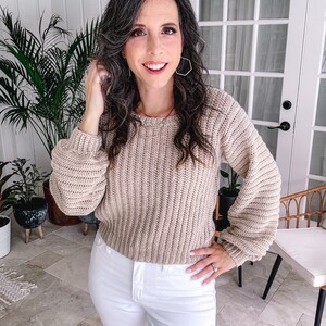 Woman posing with hand in hair, wearing a beige crochet sweater and unique jewellery, in a room with plants and French doors.