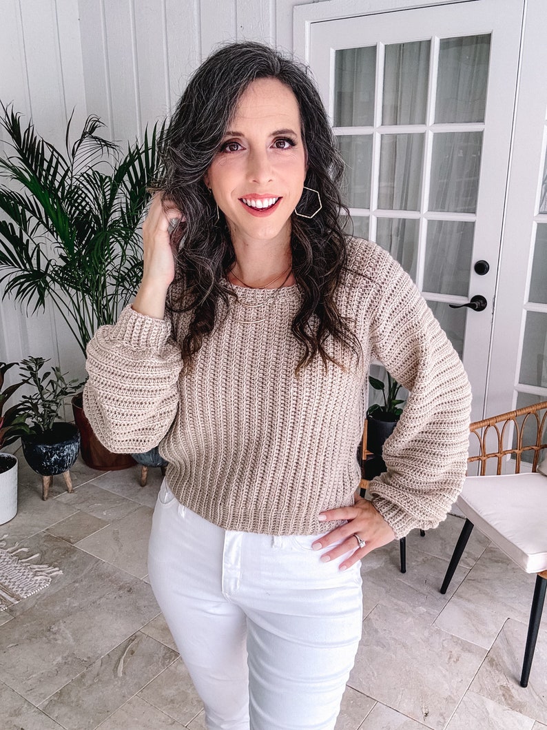 Woman posing with hand in hair, wearing a beige crochet sweater and unique jewellery, in a room with plants and French doors.