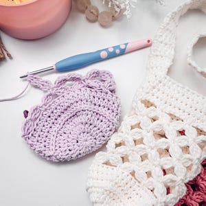 Puede incluir: Una pieza circular de ganchillo color violeta claro con borde festoneado, un bolso de ganchillo blanco con patr&oacute;n de rombos y un ganchillo azul y rosa. El ganchillo tiene un mango azul claro con lunares rosas y punta de metal plateado.