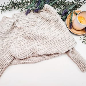 A beige crocheted sweater scarf is folded on a surface, surrounded by sprigs of lavender and a lit candle in a glass holder on a wooden tray.
