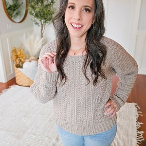 A woman with long dark hair stands in a room with wooden flooring, wearing a beige knit sweater and blue jeans. She showcases unique jewellery that highlights her look. Behind her, a round mirror and a basket with dried plants.