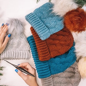 May include: A stack of five crocheted hats in various colors, including gray, blue, and orange. The hats have fluffy pom-poms on top. A crochet hook is lying on a white surface.