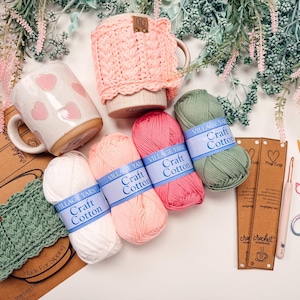May include: A collection of crochet supplies and accessories, including yarn, mugs, and crochet hooks. The image features several skeins of Village Yarn Craft Cotton in various colors, along with a pink crocheted mug cozy and a mug with pink hearts.