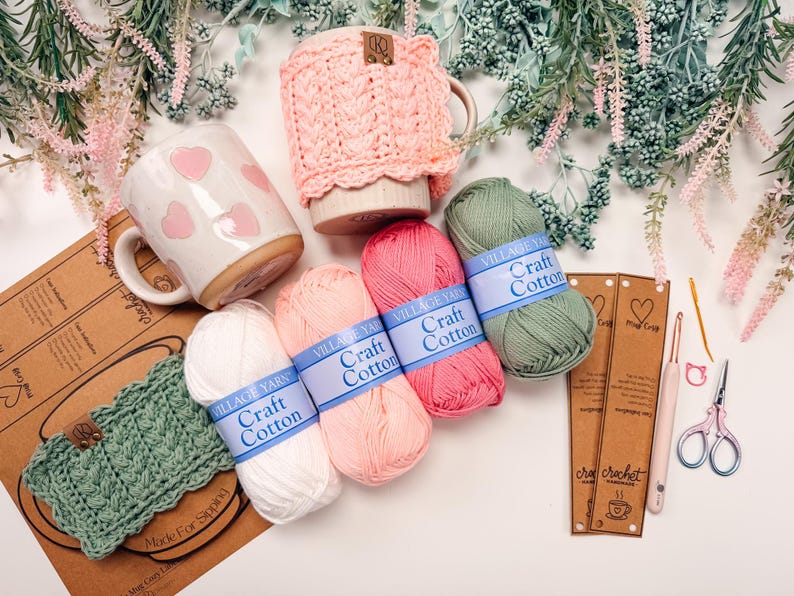 May include: A collection of crochet supplies and accessories, including yarn, mugs, and crochet hooks. The image features several skeins of Village Yarn Craft Cotton in various colors, along with a pink crocheted mug cozy and a mug with pink hearts.