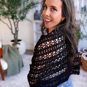 A woman with long gray hair, wearing a white top and blue jeans, holds up a large black crocheted shawl indoors—a perfect handmade gift—surrounded by plants and baskets in the background.