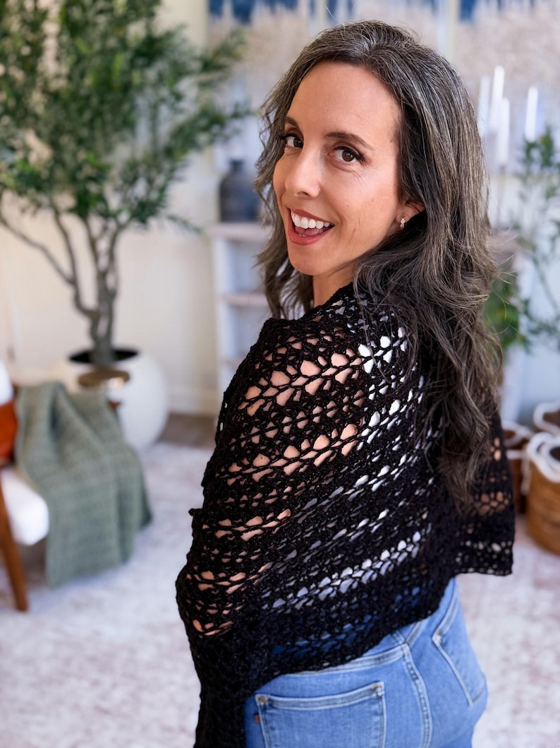 A woman with long gray hair, wearing a white top and blue jeans, holds up a large black crocheted shawl indoors—a perfect handmade gift—surrounded by plants and baskets in the background.