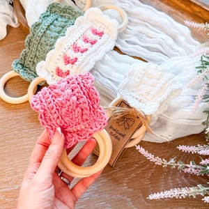 May include: A collection of handmade crochet dish towel holders in various colors, including pink, white, and green. Each holder features a wooden ring and decorative crochet patterns. The holders are displayed on a wooden tray with floral accents.