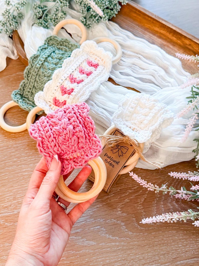 May include: A collection of handmade crochet dish towel holders in various colors, including pink, white, and green. Each holder features a wooden ring and decorative crochet patterns. The holders are displayed on a wooden tray with floral accents.