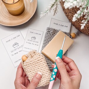 Puede incluir: Dos jaboneras de ganchillo, una beige y otra gris, con una pastilla de jabón dentro de la beige. Las jaboneras están hechas de fibra de algodón y están etiquetadas como "Scrub-A-Dub-Dub Soap Saver".