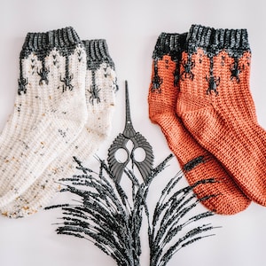 以下が含まれることがあります： クモの巣のデザインが施されたニットソックスが2足。1足は黒のクモがついた白、もう1足は黒のクモがついたオレンジ。ソックスは白い背景に黒い装飾的な枝と一緒に置かれています。