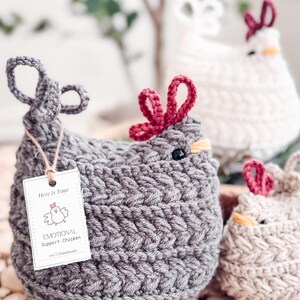 Gray crochet chicken with red comb and an emotional support chicken tag, with white and oatmeal hens behind