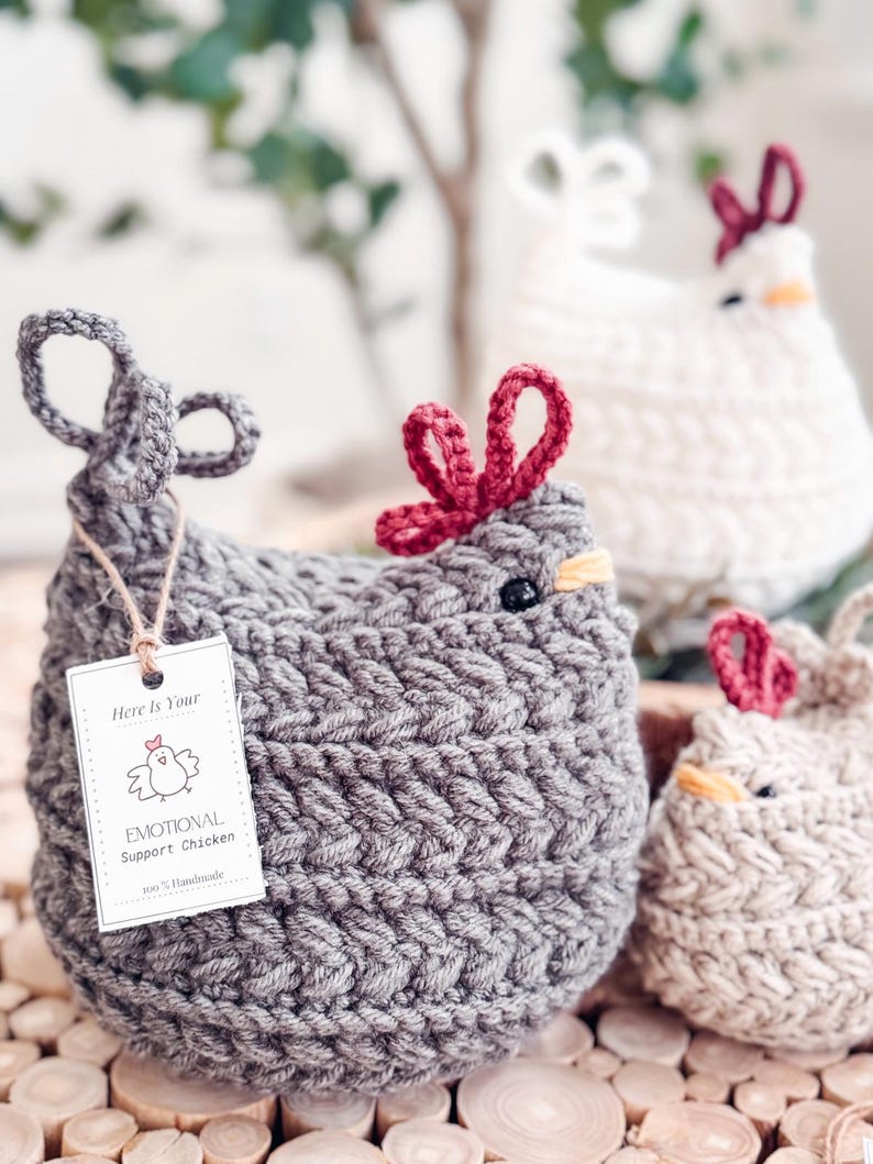 Gray crochet chicken with red comb and an emotional support chicken tag, with white and oatmeal hens behind