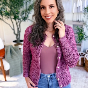 May include: A magenta crochet cardigan with pearl buttons is worn over a mauve top and blue jeans. The cardigan has a textured stitch pattern. The woman is smiling, with long gray hair.