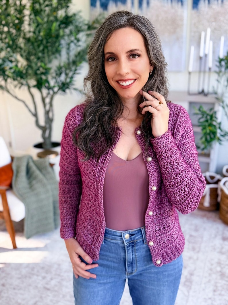 May include: A magenta crochet cardigan with pearl buttons is worn over a mauve top and blue jeans. The cardigan has a textured stitch pattern. The woman is smiling, with long gray hair.