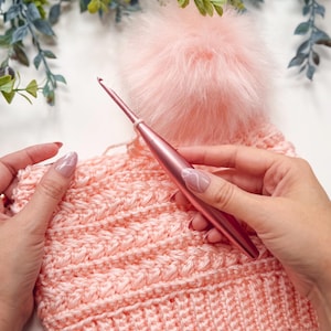 Hands holding a pink crochet hook and a partially crocheted pink hat with a fluffy pom-pom—an ideal handmade gift—set against green foliage in the background.