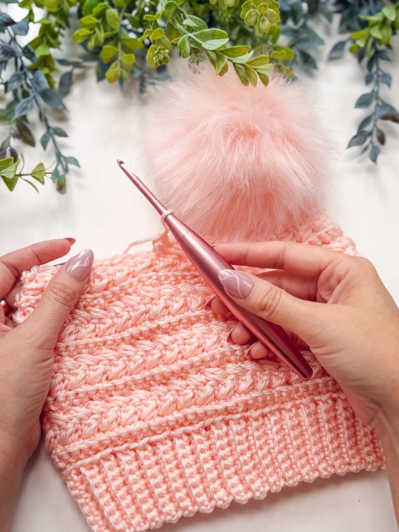 Hands holding a pink crochet hook and a partially crocheted pink hat with a fluffy pom-pom—an ideal handmade gift—set against green foliage in the background.