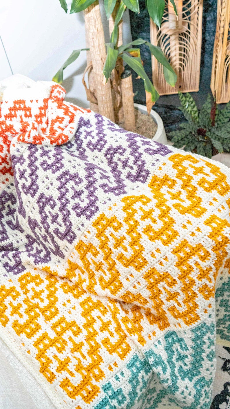Puede incluir: Una manta de ganchillo colorida con un patr&oacute;n geom&eacute;trico en tonos naranja, morado, amarillo y turquesa.