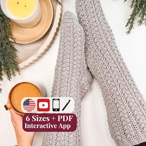 May include: A pair of light gray crocheted socks featuring a vertical braided cable pattern. An overlay graphic includes the text 6 Sizes + PDF Interactive App, along with icons for the American flag, a YouTube play button, a smartphone, and a crochet hook.