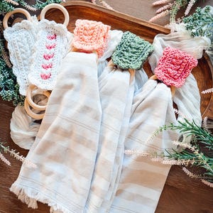 May include: A collection of decorative kitchen towels with crocheted ring holders. The towels are beige with horizontal stripes and fringed edges. The ring holders are in various colors, including white, pink, and green, and are made of crocheted yarn and wooden rings.