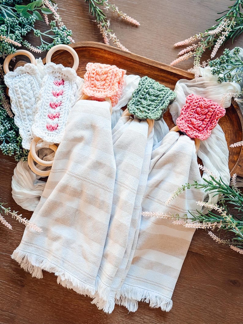 May include: A collection of decorative kitchen towels with crocheted ring holders. The towels are beige with horizontal stripes and fringed edges. The ring holders are in various colors, including white, pink, and green, and are made of crocheted yarn and wooden rings.