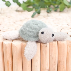 May include: A plush turtle toy with a light green shell and light gray flippers and head. The turtle has black button eyes and is resting on a light brown wooden surface. The background includes green foliage.