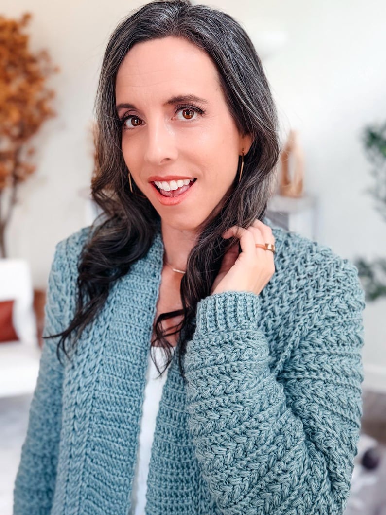 May include: A light blue crochet cardigan with a textured stitch pattern. The open-front sweater has long sleeves and a relaxed fit. The cardigan is worn over a white top.