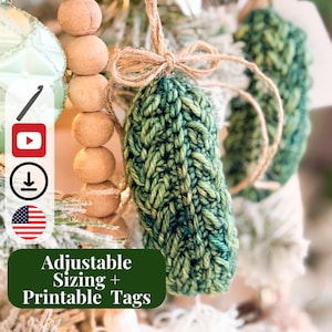 May include: A green crocheted pickle ornament hanging from a jute twine bow. The ornament features a textured yarn stitch pattern. A dark green rectangular label with white text reads Adjustable Sizing + Printable Tags, alongside icons for crochet, video, download, and the American flag.