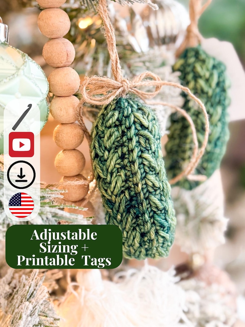 May include: A green crocheted pickle ornament hanging from a jute twine bow. The ornament features a textured yarn stitch pattern. A dark green rectangular label with white text reads Adjustable Sizing + Printable Tags, alongside icons for crochet, video, download, and the American flag.