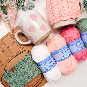 May include: A white ceramic mug with pink hearts, a pink crocheted mug cozy, and a green crocheted mug cozy. Several skeins of Village Yarn Craft Cotton in white, pink, and green are also present. The image also includes a printed pattern.