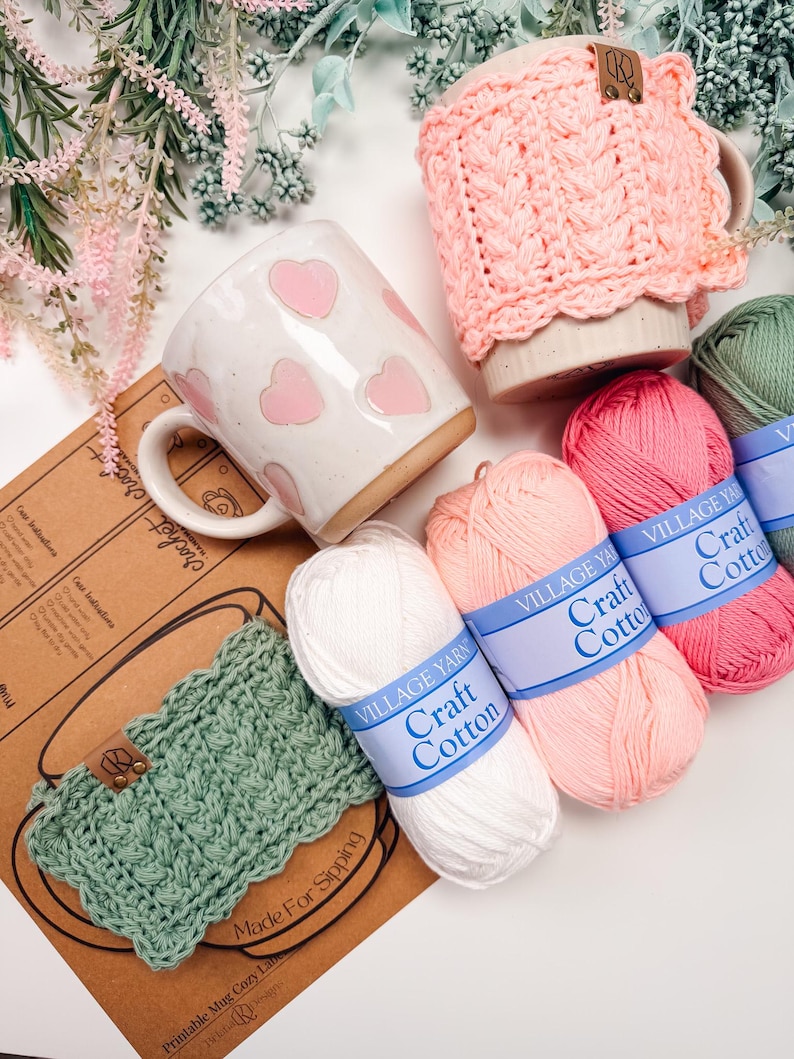 May include: A white ceramic mug with pink hearts, a pink crocheted mug cozy, and a green crocheted mug cozy. Several skeins of Village Yarn Craft Cotton in white, pink, and green are also present. The image also includes a printed pattern.