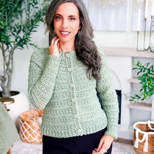 May include: A light green crochet cardigan with a button-up front. The cardigan has a textured stitch pattern and long sleeves. The buttons are a matching light green color. The person is wearing black pants.